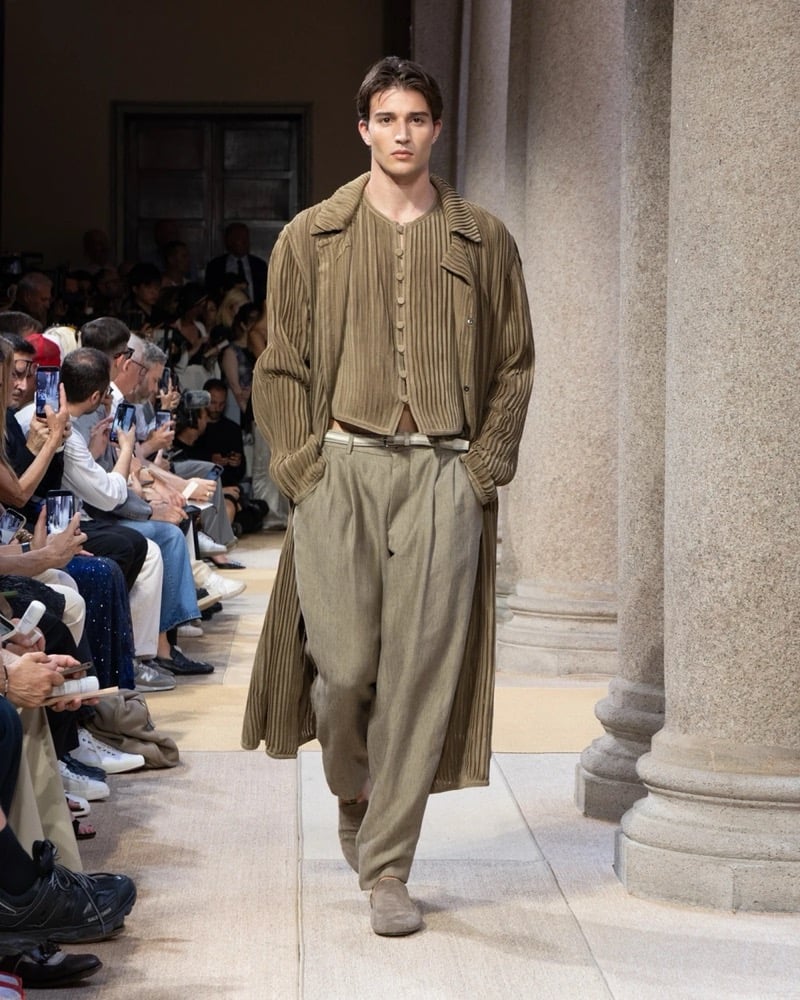 Model wears a layered ribbed-pleat duster coat over a cropped ribbed cardigan, paired with wide pleated drawstring linen trousers