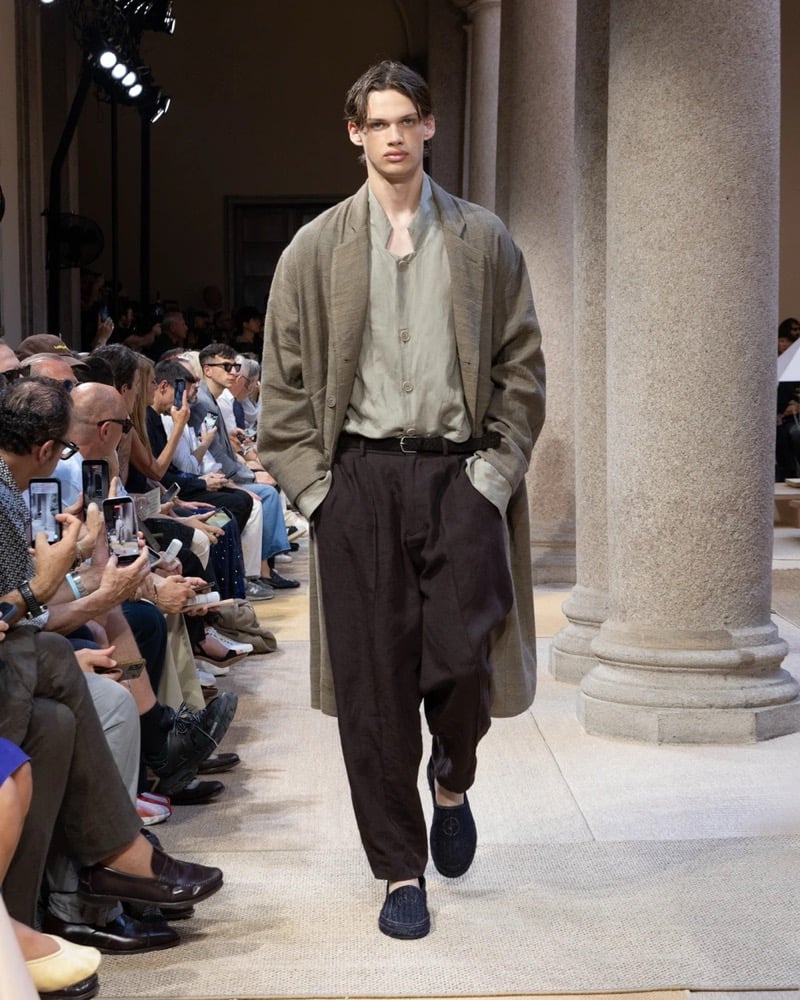 Model wears an oversized notch-lapel linen overcoat layered over a half-buttoned shirt, paired with wide pleated linen trousers