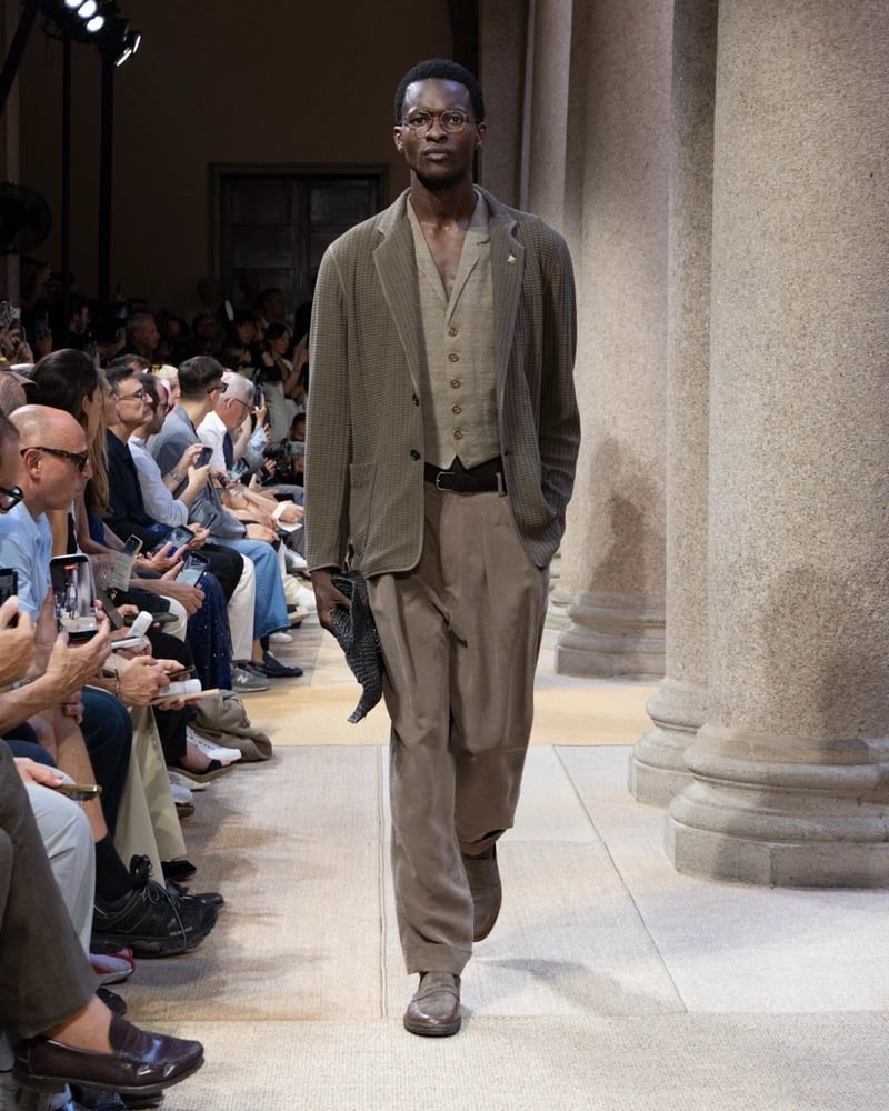 Model wears a single-button linen blazer with notch lapels layered over a buttoned waistcoat and open shirt, paired with wide pleated linen trousers
