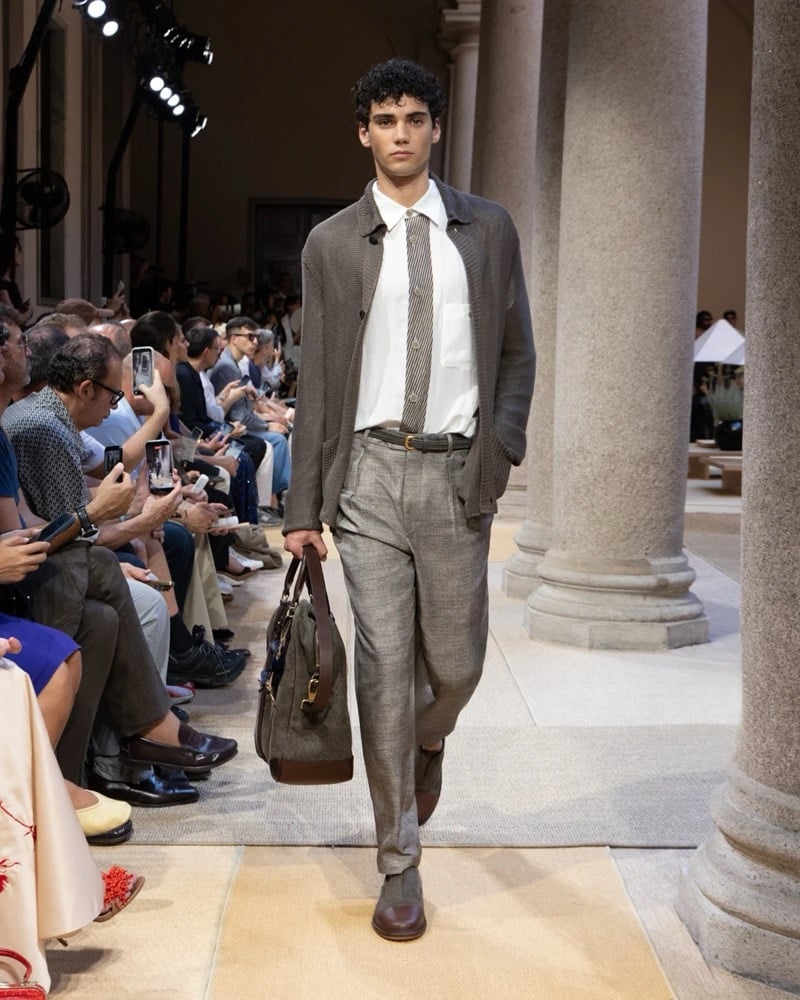 Model wears a relaxed linen shirt-jacket layered over a shirt and knit tie, paired with wide pleated chevron linen trousers