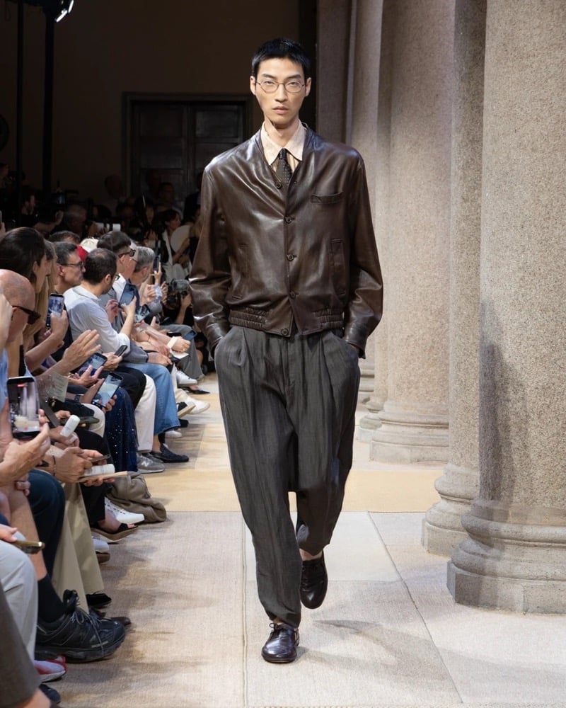 Model wears a soft leather blouson with a band collar and chest flap pocket over an open shirt, paired with wide pleated pinstripe trousers