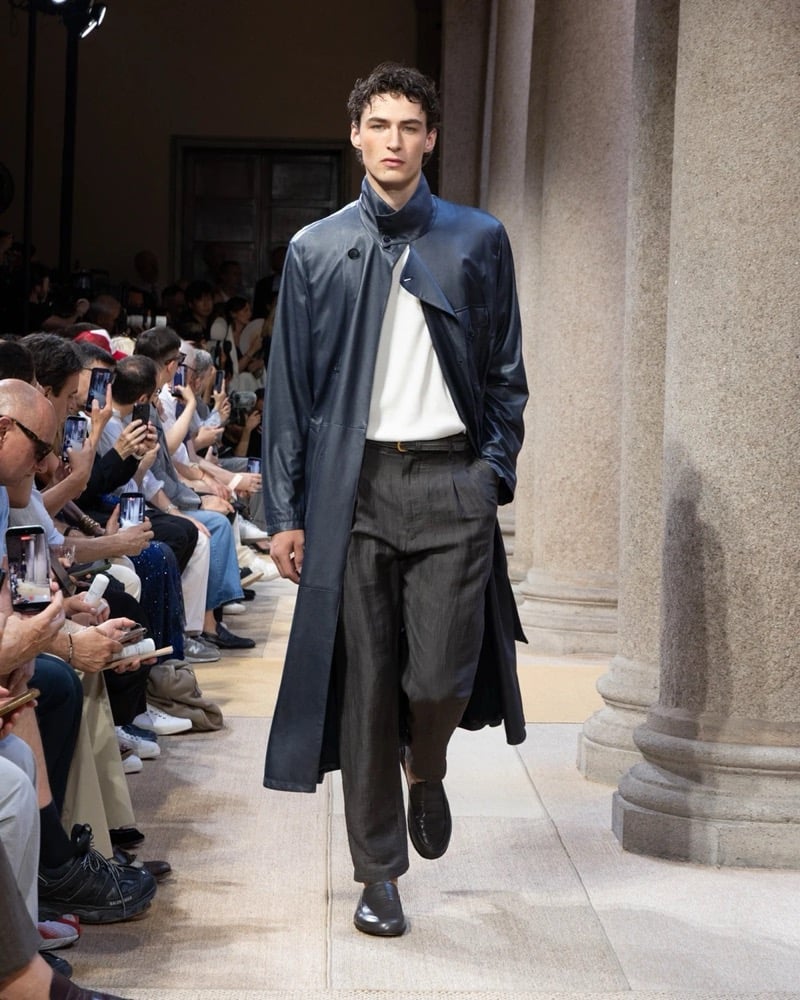 Model wears a long stand-collar leather coat with a half-button placket layered over a tunic, paired with wide pleated linen trousers