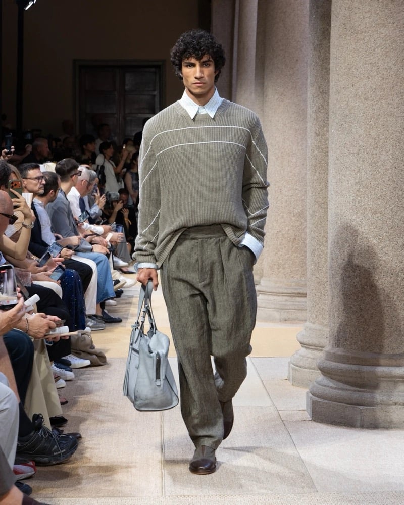 Model wears a ribbed crewneck knit pullover with a horizontal stripe across the chest, layered over a soft shirt with wide pleated linen trousers