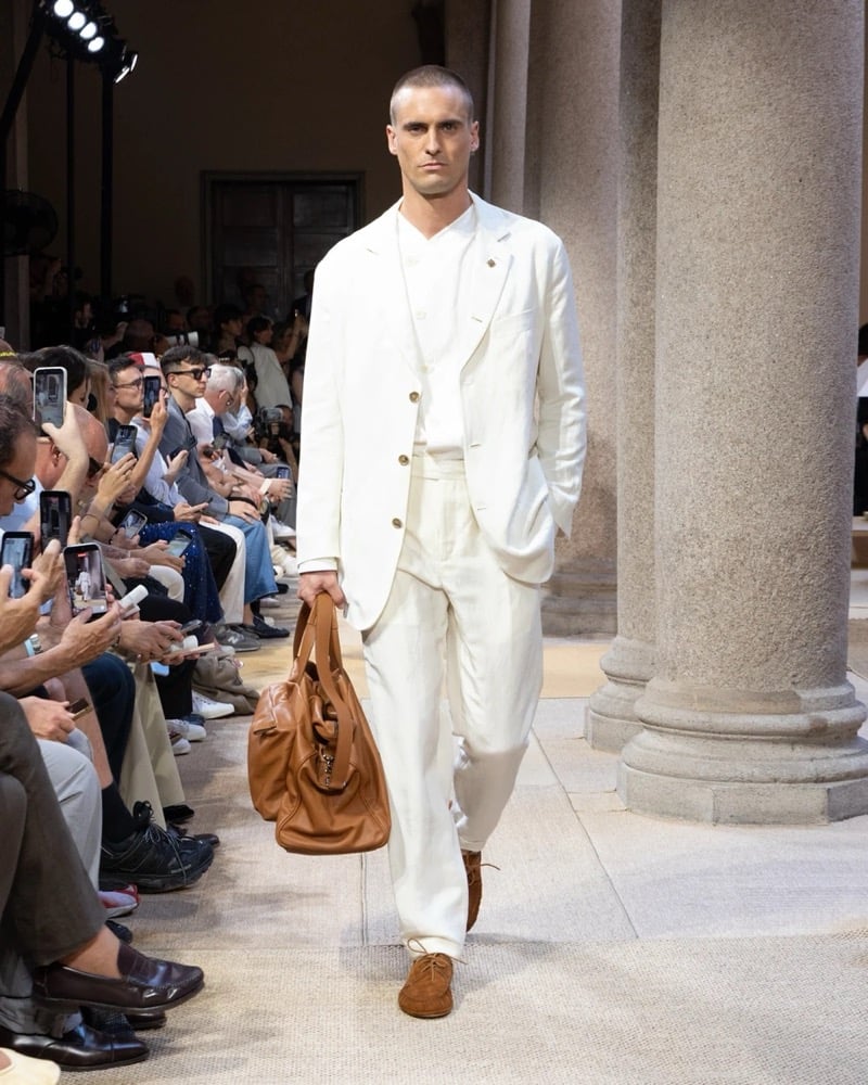 Model wears a relaxed single-button linen blazer with notch lapels layered over a buttoned waistcoat, paired with wide pleated linen trousers
