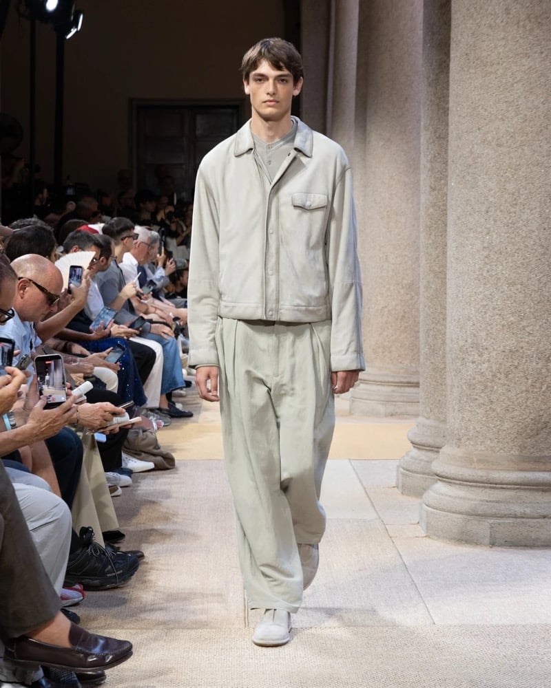 Model wears a relaxed suede shirt-jacket with a zip front and a chest flap pocket layered over a polo, paired with wide pleated linen trousers