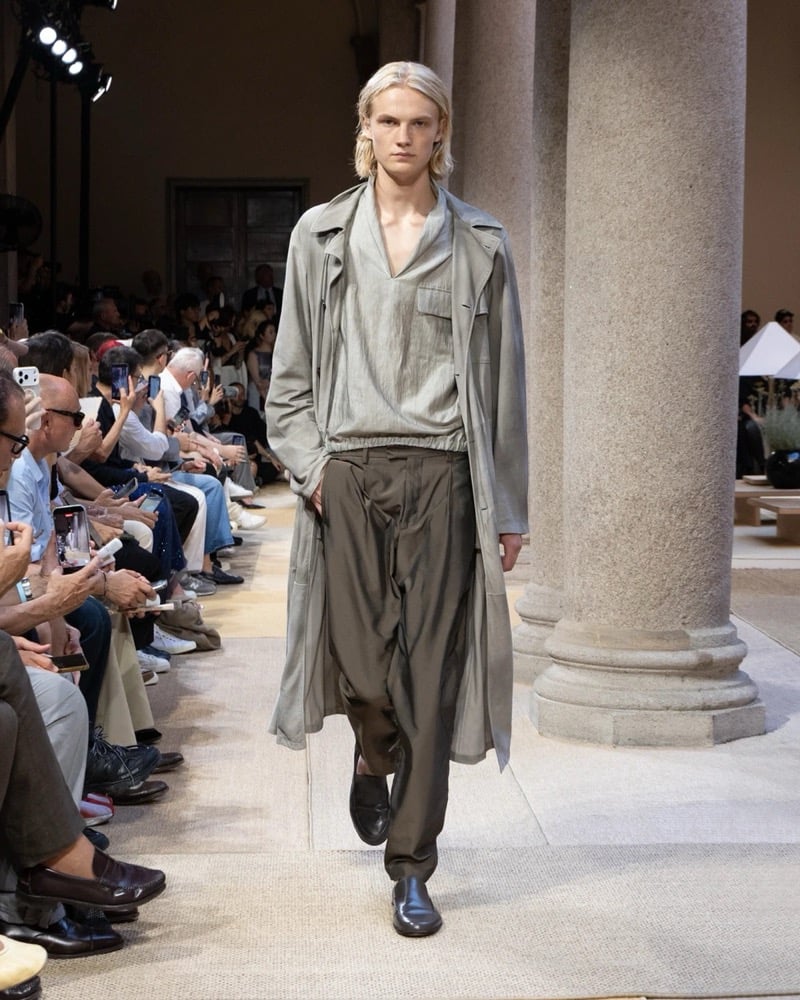 Model wears a relaxed long open trench coat layered over a wrap-front tunic, paired with wide pleated drawstring trousers