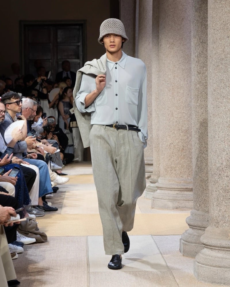 Model wears a long-sleeve button-up shirt with a chest patch pocket paired with wide pleated linen trousers, a folded jacket carried over the shoulder