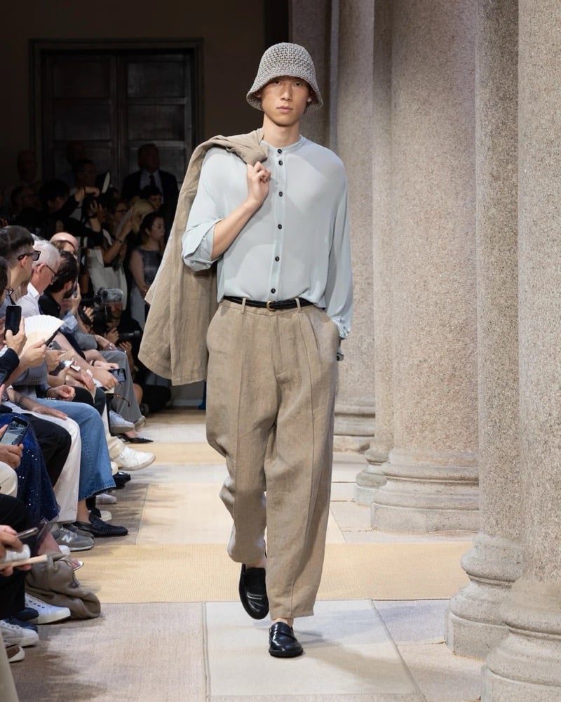 Model wears a band-collar long-sleeve sheer button-up shirt paired with wide pleated linen trousers, a folded linen jacket carried over the shoulder