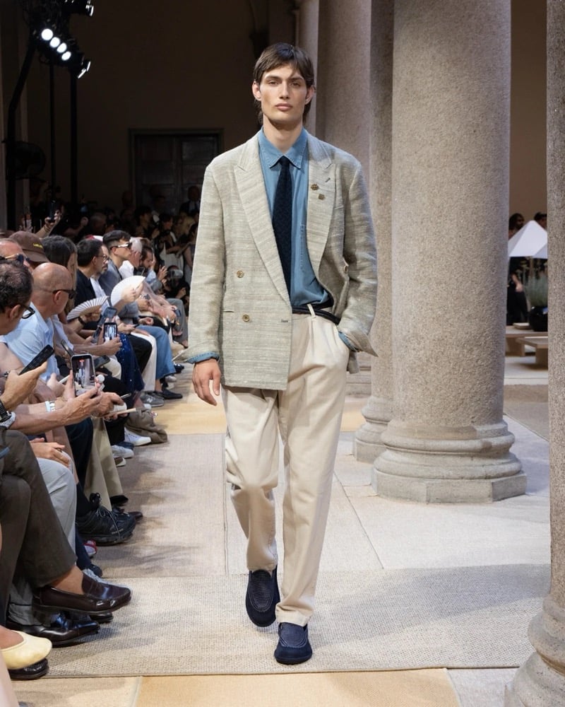 Model wears a double-breasted textured linen blazer with notch lapels layered over a shirt and patterned tie, paired with wide pleated trousers