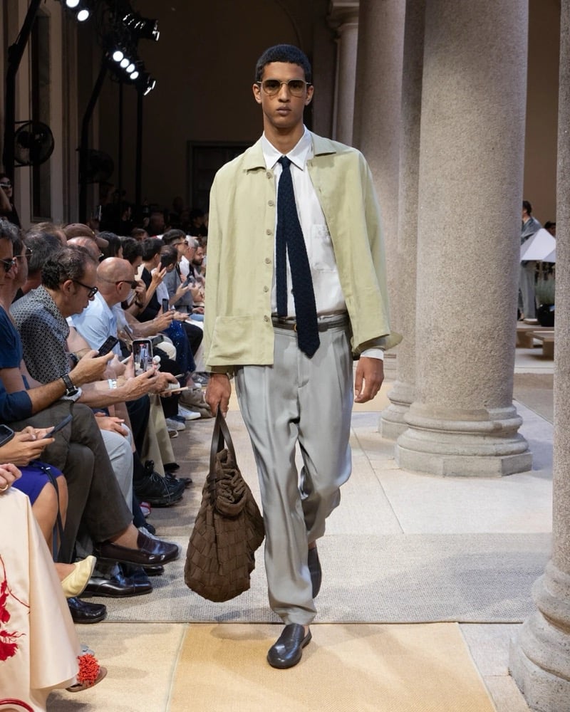 Model wears a relaxed silk-blend shirt-jacket with a button front layered over a shirt and patterned tie, paired with wide pleated silk trousers