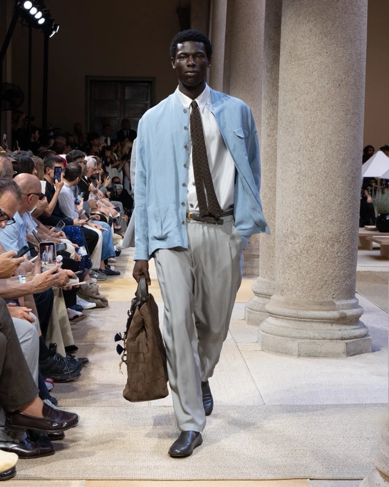 Model wears a soft linen shirt-jacket with a chest flap pocket layered over a shirt and patterned tie, paired with wide pleated silk trousers