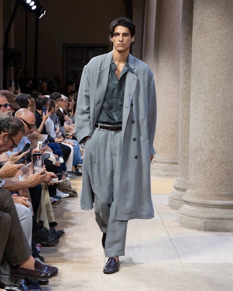 Model wears a long open linen trench coat layered over a camp-collar shirt, paired with wide pleated linen trousers