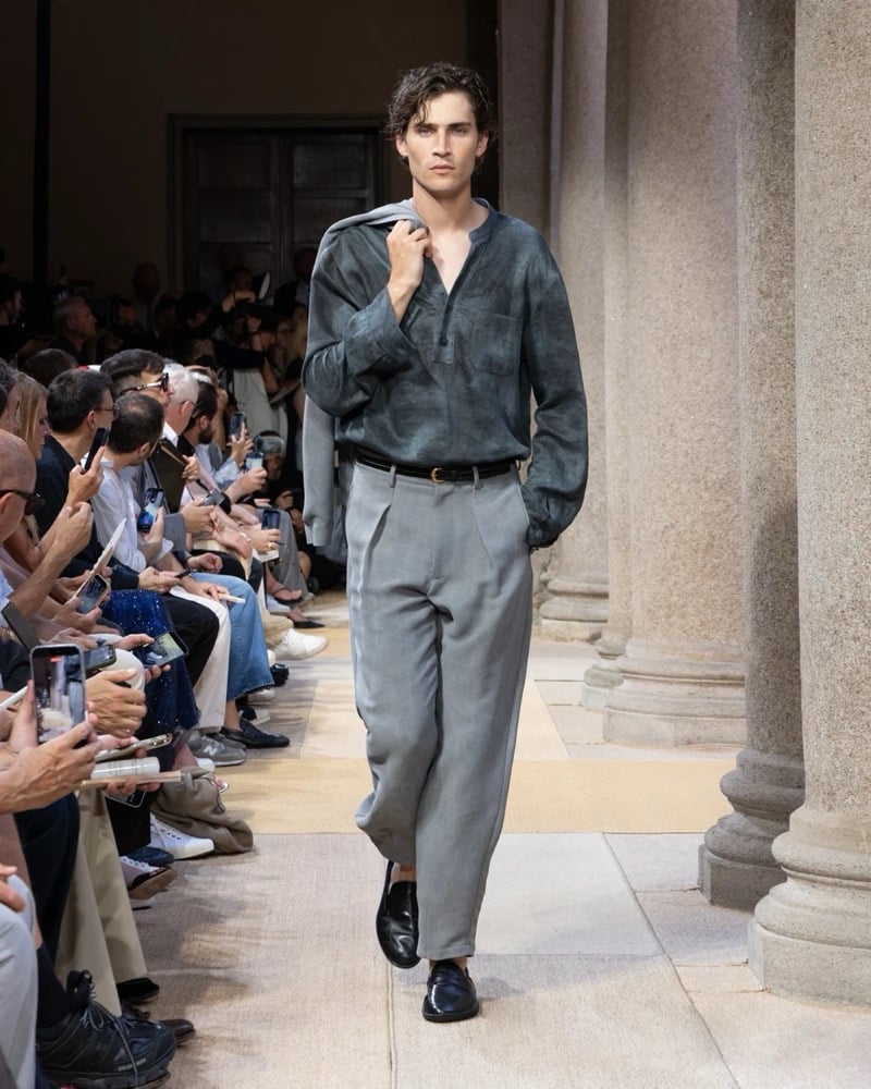 Model wears a long-sleeve linen tunic with a half-button placket and chest patch pocket, paired with wide pleated linen trousers, a jacket carried over the shoulder