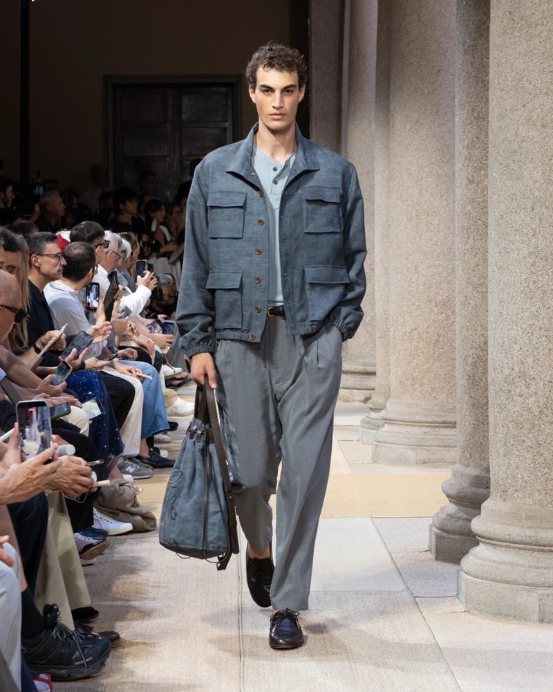 Model wears a linen safari jacket with four flap pockets and a banded hem layered over a polo, paired with wide pleated linen trousers