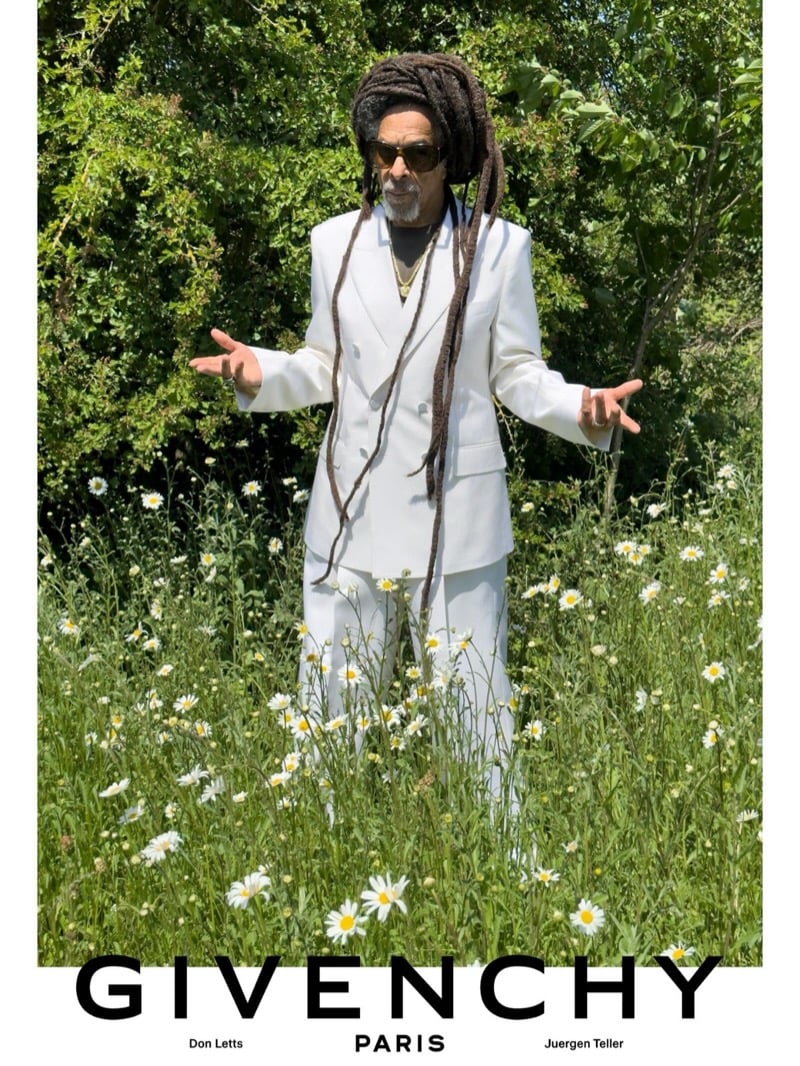 Don Letts opens his arms in a white Givenchy double-breasted suit, sunglasses, and a gold pendant chain while standing in a meadow of daisies