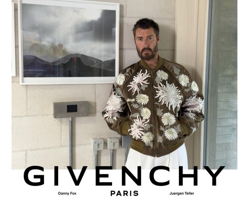 Danny Fox wears a Givenchy bomber jacket embroidered with chrysanthemums, paired with white wide-leg trousers, posed beside a framed volcano photograph