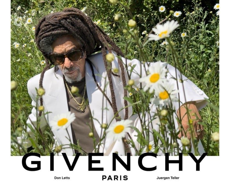 Don Letts relaxes in a wildflower meadow in a white Givenchy suit jacket layered over a knit tee, finished with a gold pendant chain and tortoise sunglasses
