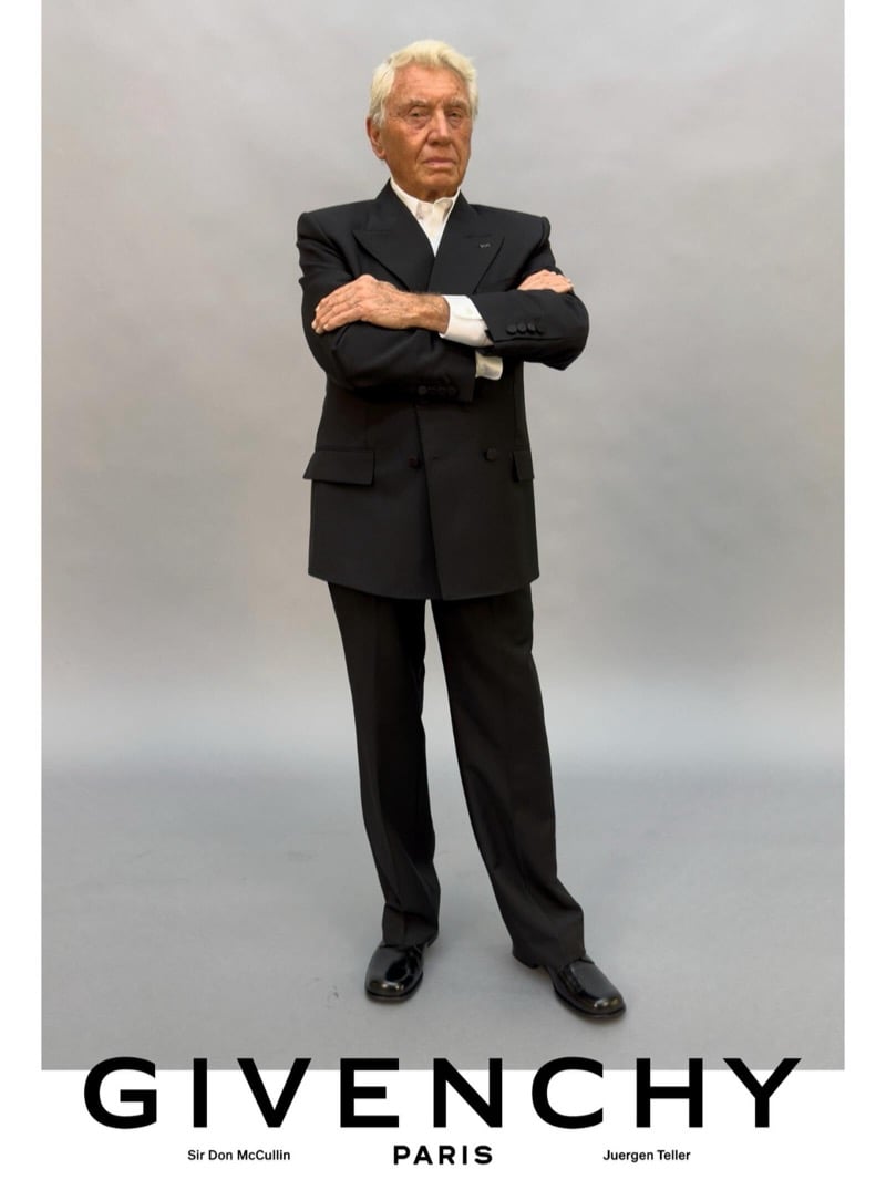 Sir Don McCullin crosses his arms in a Givenchy double-breasted suit with an open-collar dress shirt and polished leather oxfords against a gray studio backdrop