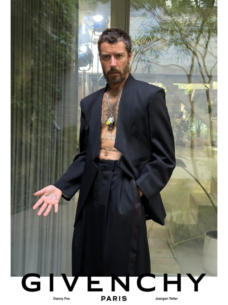 Danny Fox opens his jacket to reveal a tattooed torso and silver pendant necklace, wearing a Givenchy tailored suit with wide pleated trousers