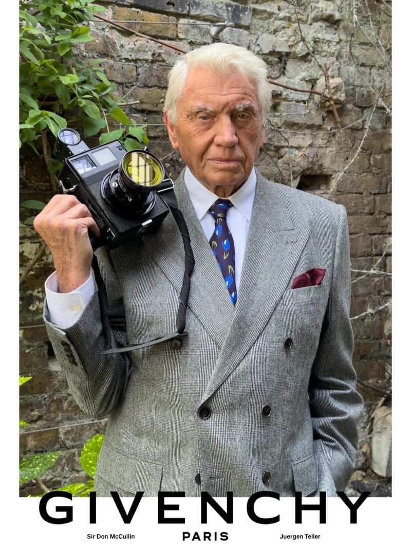 Sir Don McCullin rests a vintage rangefinder camera on his shoulder in a Givenchy glen check double-breasted suit, silk tie, and burgundy pocket square