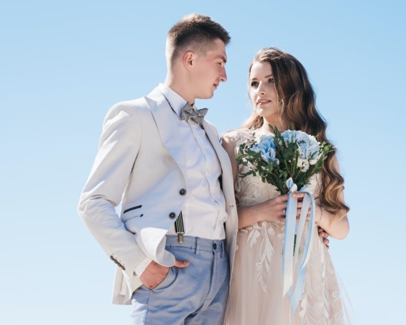 Groom in a light blazer, a lighter vest, mid-blue trousers, and a bow tie standing beside his bride in a pale lace gown holding a bouquet of blue flowers under an open sky