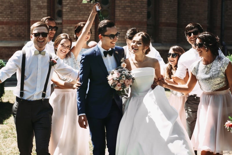 Groom in a blue suit and a bow tie walking with his bride in a strapless white gown carrying a pastel bouquet, surrounded by their wedding party laughing in pale dresses and shirtsleeves with suspenders