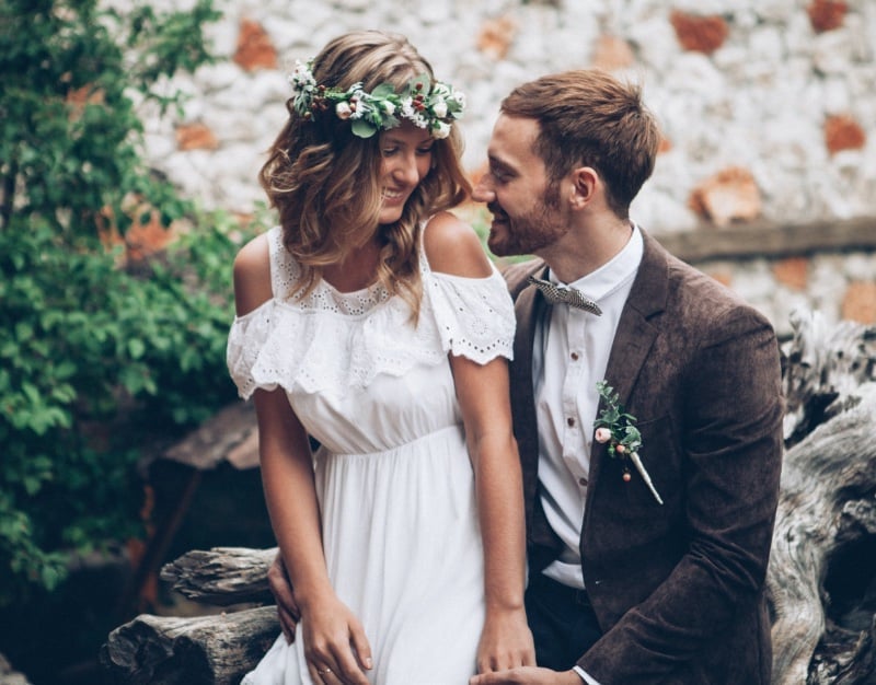 Groom in a textured brown jacket and a small printed bow tie with a green boutonniere leaning toward his bride in an off-shoulder eyelet dress and a flower crown, seated on driftwood against a stone wall