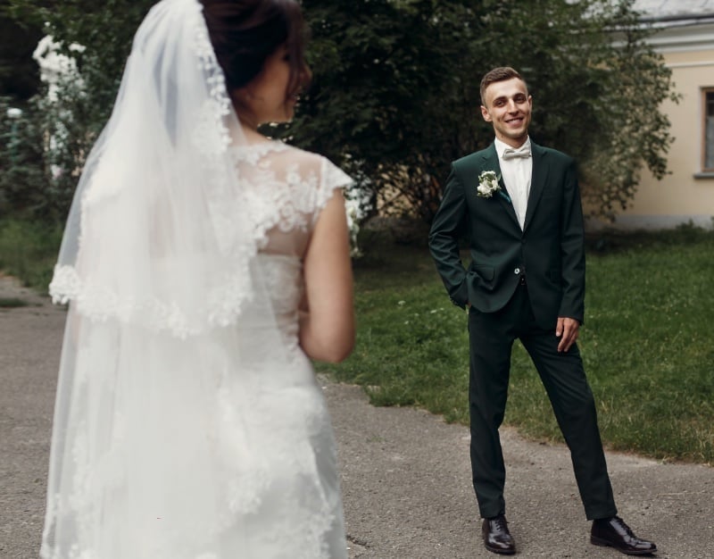 Groom in a dark green two-piece suit with a small boutonniere standing on a stone path as his bride in a lace gown and long veil sees him for the first time, framed soft in the foreground