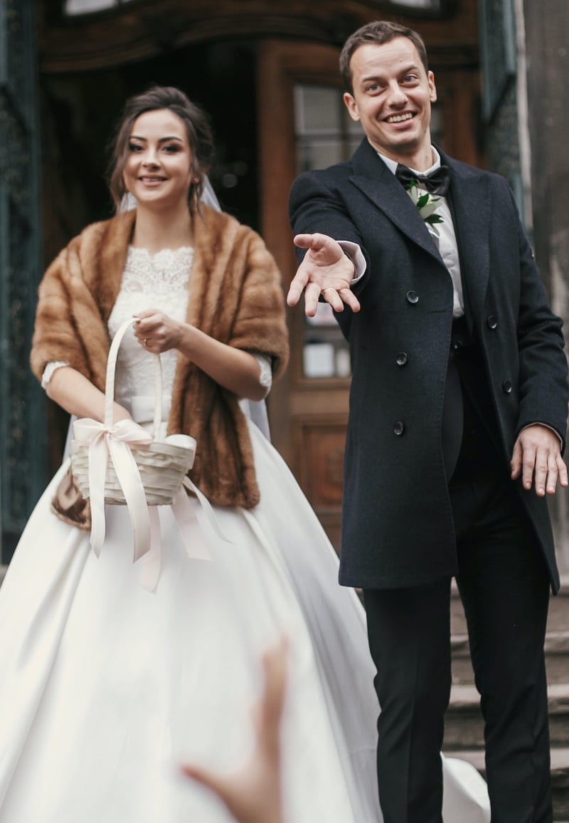 Groom in a long dark overcoat and a bow tie reaching out a hand on a stone church step, his bride beside him in a full white ball gown with a tan fur stole holding a small basket