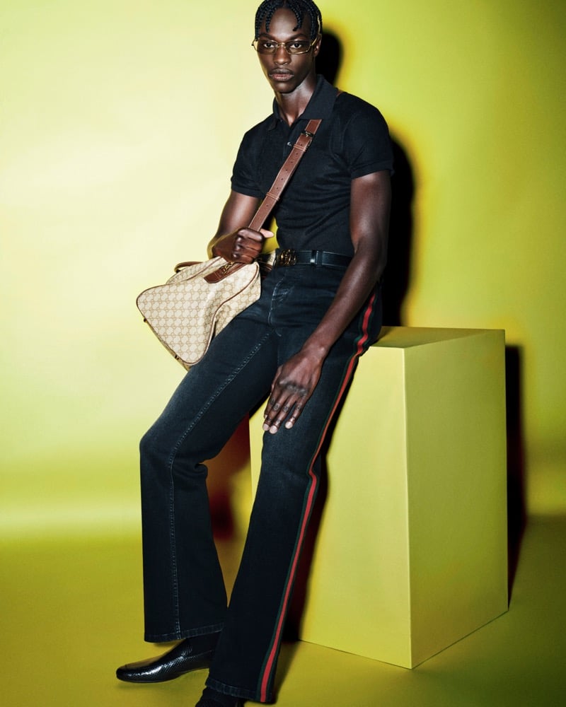 Abdou Thior leaning against a yellow plinth in a black short-sleeve polo and slim rectangular sunglasses, dark jeans striped vertically in red and green down each leg, a GG-monogram canvas tote with brown leather trim and Web stripe slung over the shoulder