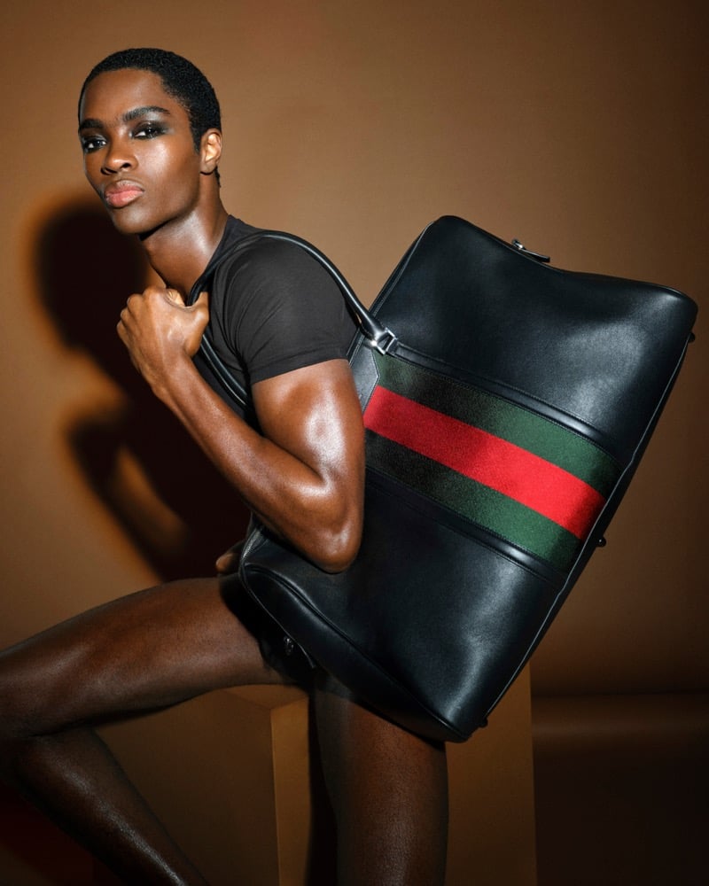 Alton Mason in a fitted black short-sleeve tee against a warm brown backdrop, shouldering an oversized black leather duffel boldly banded in the red-and-green Web stripe, one knee raised on a plinth