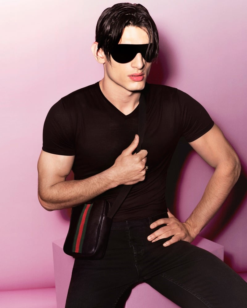 Ardi Hodzic in a fitted black short-sleeve V-neck tee and black slim trousers against a soft pink studio backdrop, dark hair falling over oversized black shield sunglasses, a black leather crossbody bag with the red-and-green Web stripe pulled tight across the chest