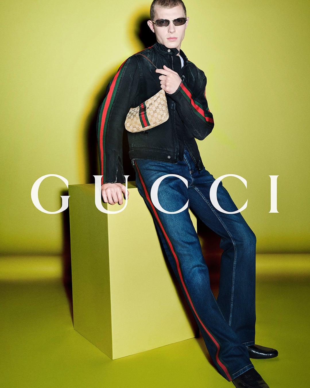 Bede Harper standing against a chartreuse backdrop printed with the white Gucci logo, wearing a black denim moto jacket detailed with red-and-green Web stripes down the sleeves and dark wash flared jeans with matching stripes down each leg, holding a small GG-monogram canvas pouch with Web stripe