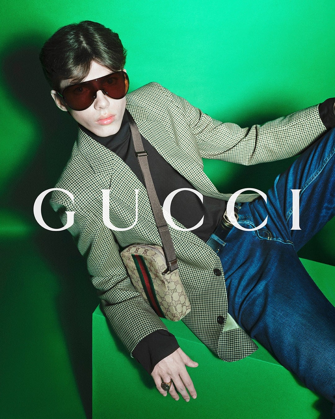 Ben Eckstein reclining across a green plinth in an oversized brown-and-white houndstooth blazer over a black turtleneck and dark wash flared jeans, a GG-monogram canvas crossbody with red-and-green Web stripe across the chest, oversized amber-tinted shield sunglasses