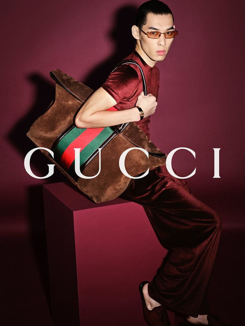 Cao Chang leaning back on a magenta plinth in a deep red velvet short-sleeve top and matching wide-leg velvet trousers, slick-back hair and slim tortoiseshell rectangular sunglasses, holding an oversized brown suede tote marked with the red-and-green Web stripe against a burgundy backdrop printed with the white Gucci logo
