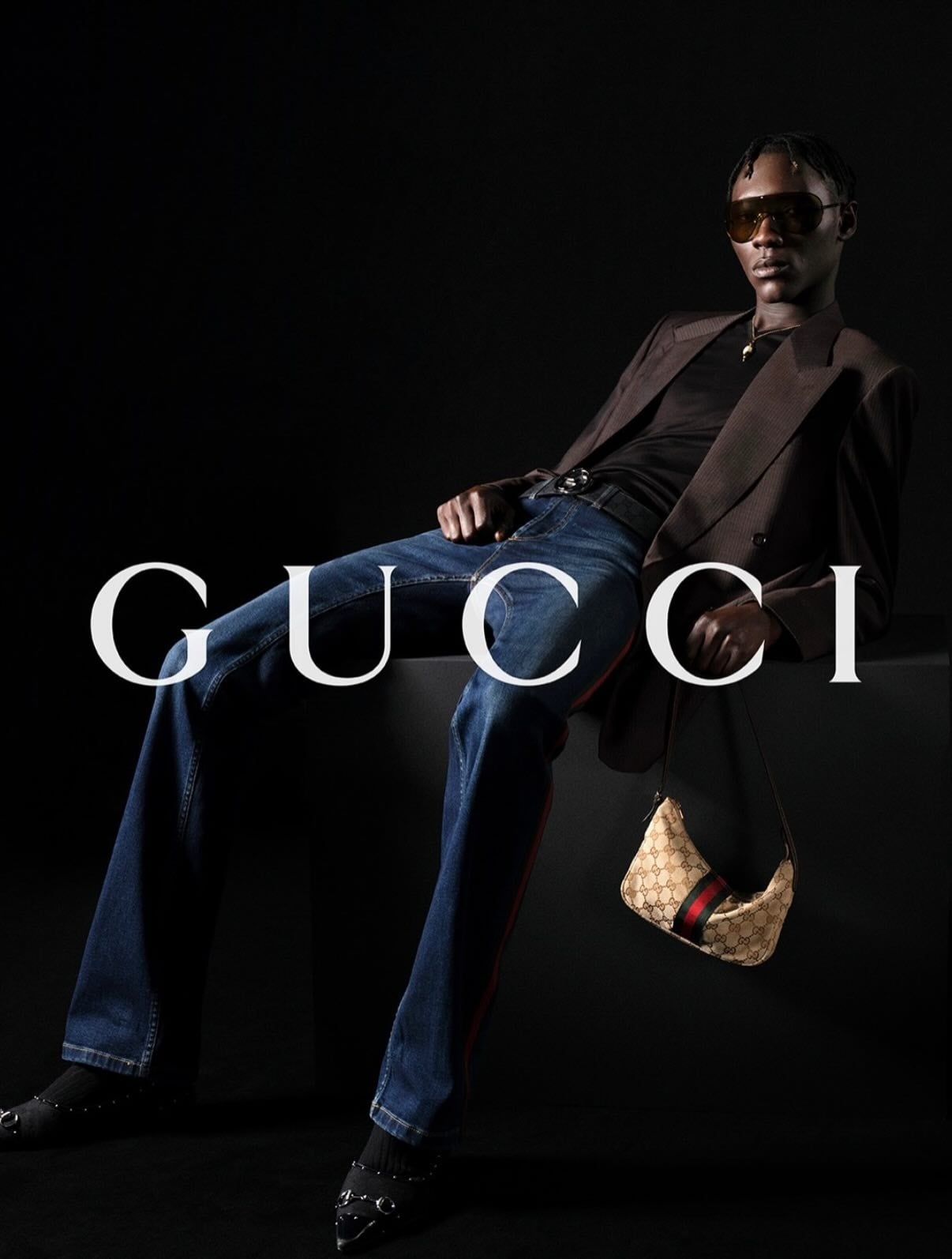 Cheikh Mara Niang seated on a low black plinth in a brown pinstripe double-breasted blazer, dark blue flared jeans striped down the leg in red, a small GG-monogram canvas hobo bag with Web stripe hanging at the hip, oversized aviator sunglasses against a near-black backdrop printed with the white Gucci logo