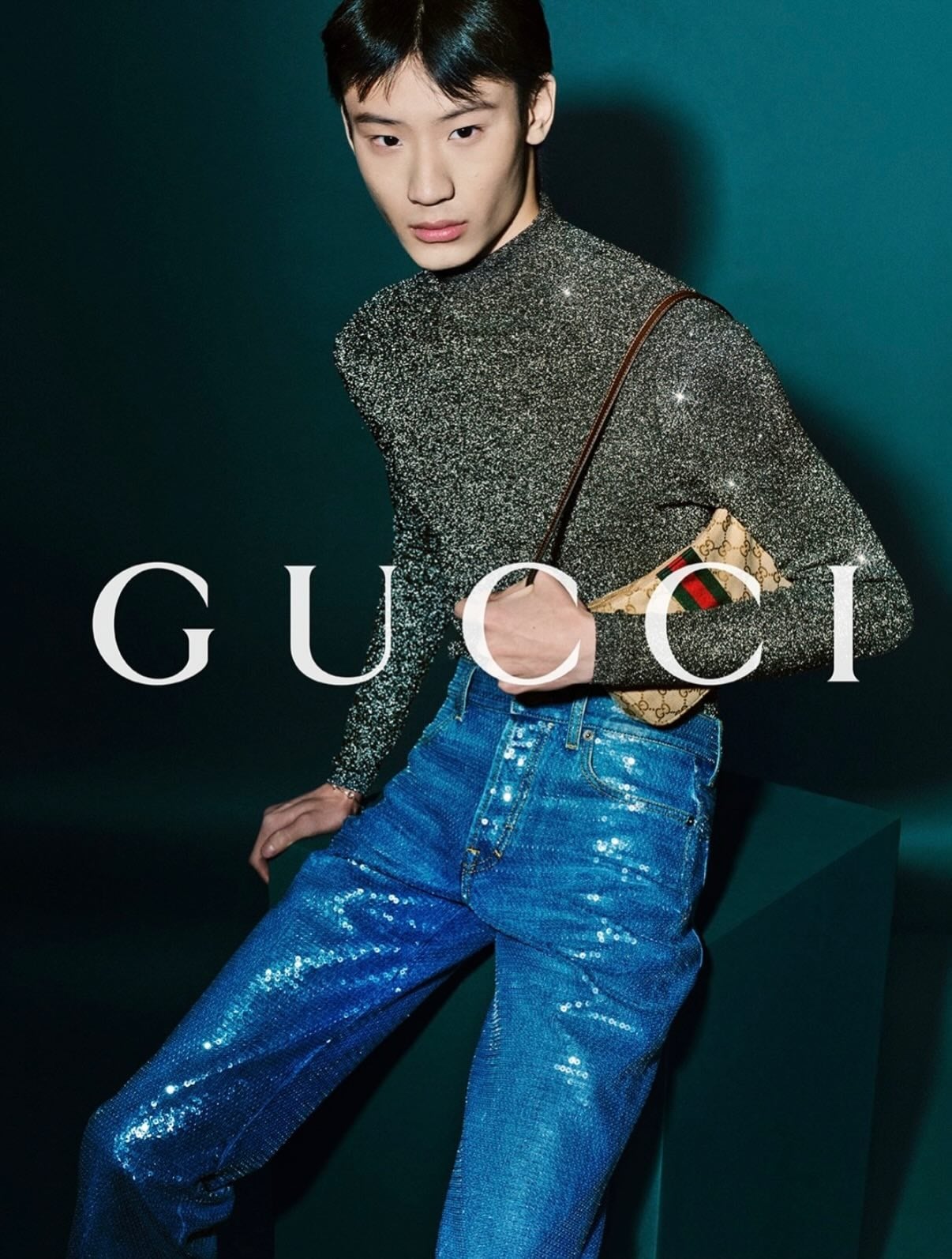 Dongliang He against a deep teal backdrop printed with the white Gucci logo, wearing a shimmering silver metallic mock-neck knit top and blue sequined jeans, a small GG-monogram canvas crossbody with Web stripe at the waist