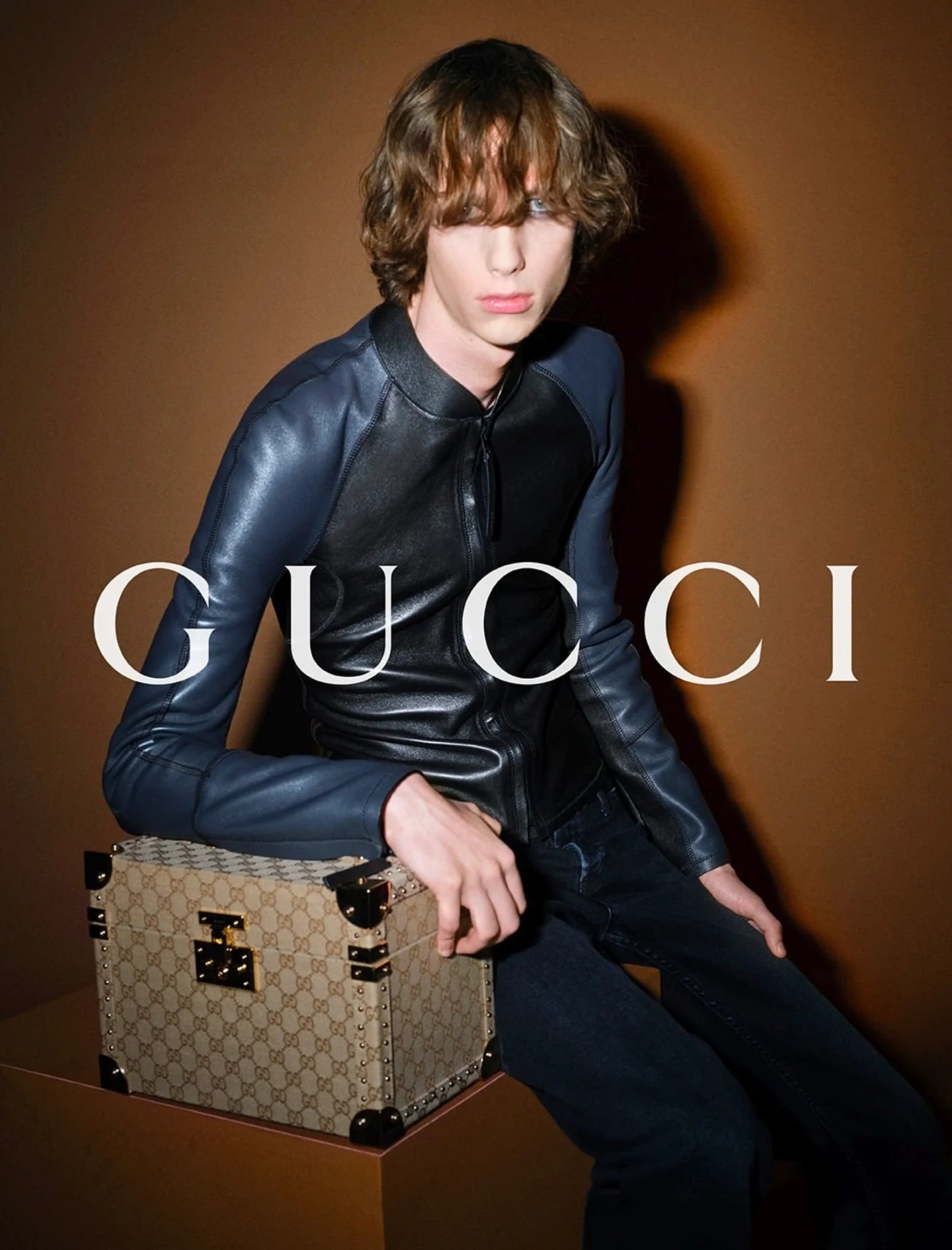 Francois Deaconu with shaggy chestnut hair, seated on a GG-monogram canvas trunk trimmed in dark leather and brass hardware, wearing a slim navy and black paneled leather zip jacket and dark jeans against a warm brown backdrop printed with the white Gucci logo