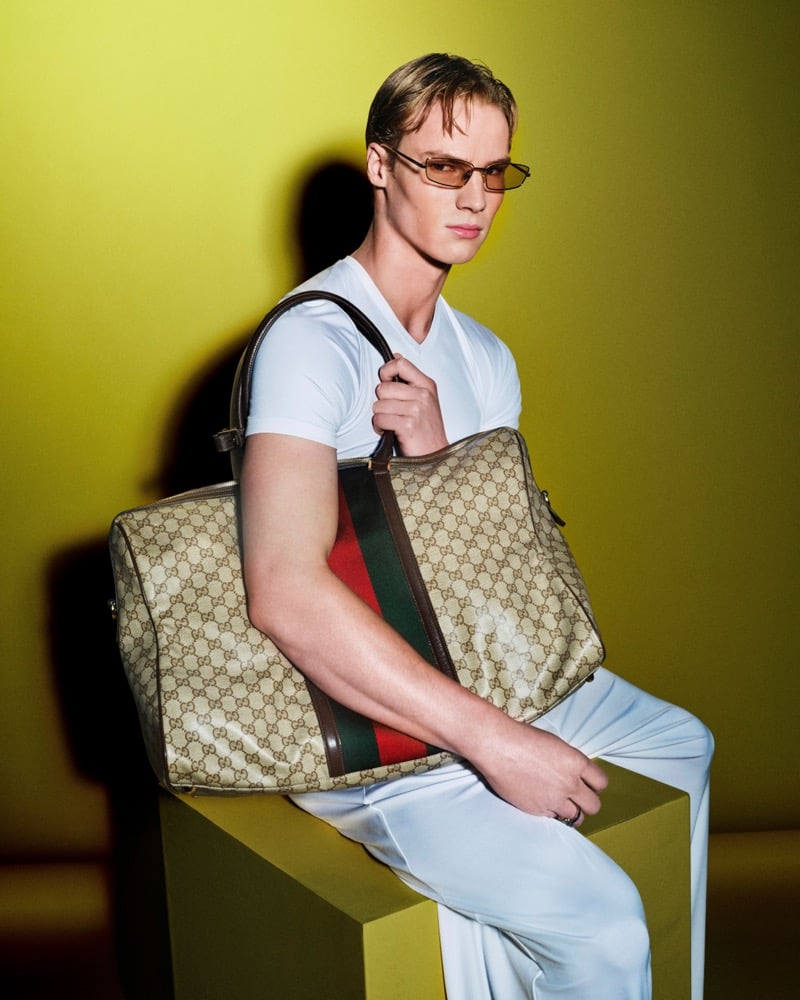 Henry Rank seated on a yellow plinth in a fitted white short-sleeve V-neck tee and pale wide-leg trousers, cradling an oversized GG-monogram canvas tote trimmed in brown leather and the red-and-green Web stripe against a chartreuse backdrop