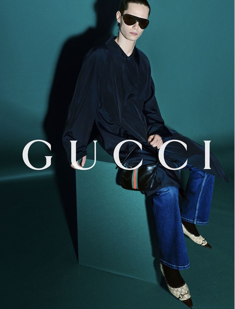 James Moody perched on a teal plinth in a fluid navy silk shirt-coat and blue wide-leg jeans, pointed-toe shoes covered in GG-monogram canvas with dark leather toes and Horsebit hardware, a small black leather shoulder bag with red-and-green Web stripe resting in the lap, oversized aviator sunglasses