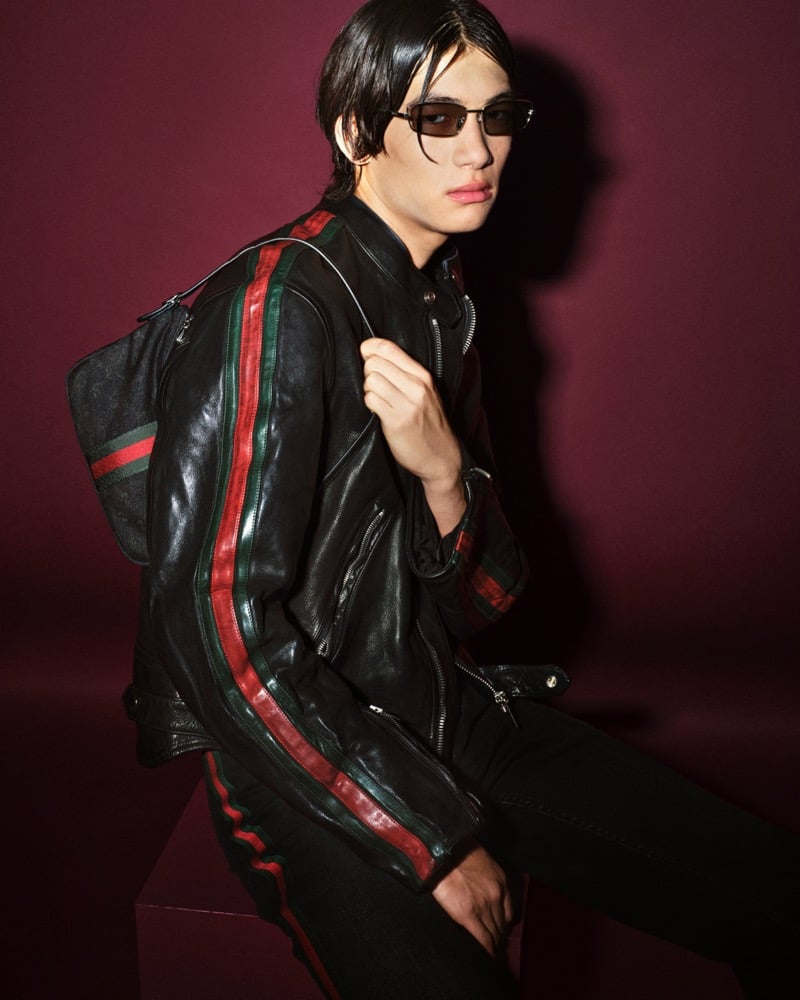 Kai Wijers seated on a low plinth against a deep burgundy backdrop, wearing a black leather moto jacket finished with vertical red-and-green Web stripes down the chest and sleeves, paired with black jeans, slim rectangular sunglasses, and a black Web-stripe backpack slung over one shoulder