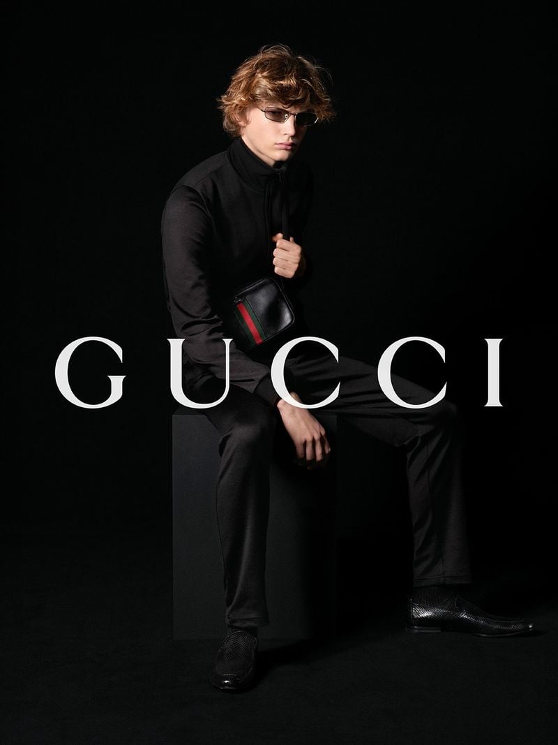 Keygon Baxter with tousled curly blond hair seated against a pitch-black backdrop in a black zip-up track jacket and slim black trousers, holding a small black leather pouch with red-and-green Web stripe at the chest, slim aviator sunglasses with metal frames