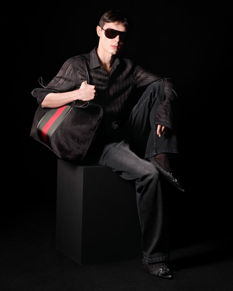 Loic Paulmier seated on a black plinth against a black backdrop in a sheer black striped shirt and dark jeans with a Horsebit-buckle belt, holding an oversized black calf-hair duffel banded in the red-and-green Web stripe, oversized black shield sunglasses covering the eyes