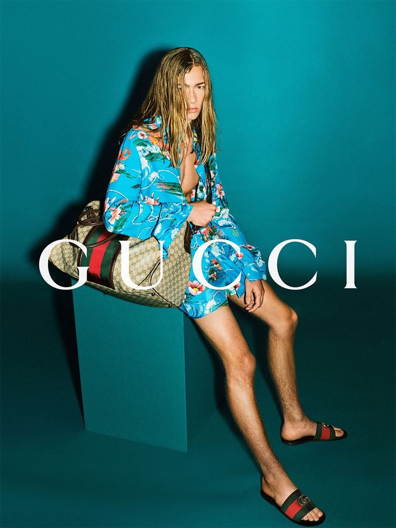 Mack Karpes with long wet blond hair seated on a teal plinth in a turquoise tropical Hawaiian-print shirt and matching shorts, GG-monogram canvas Web-stripe duffel in the lap, black-and-green Web-stripe Horsebit slides on bare feet against a deep teal backdrop
