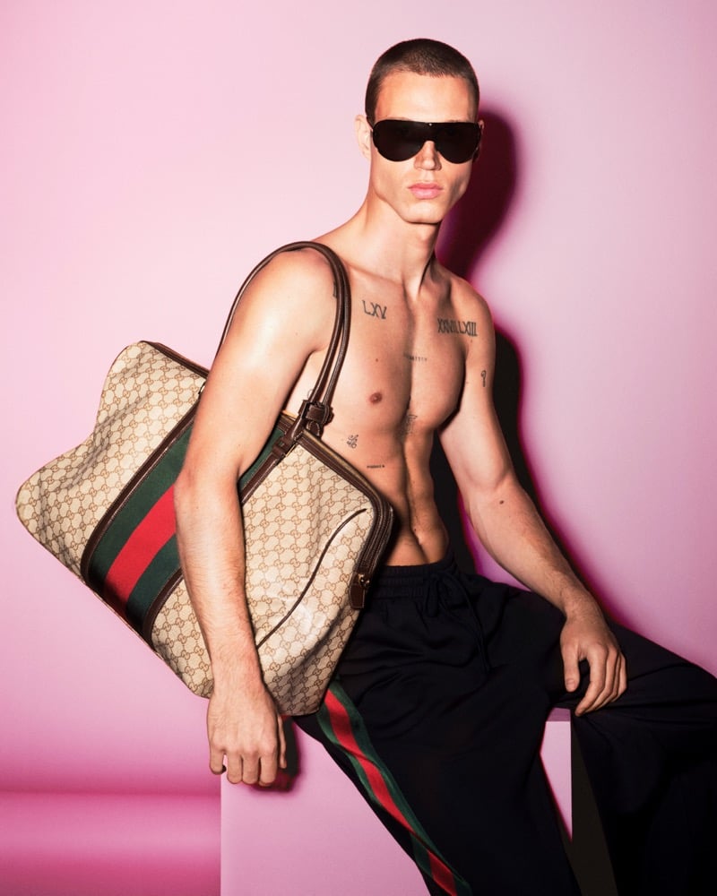 Michael Oder shirtless with a buzz cut and oversized black aviator sunglasses, Roman-numeral chest tattoos visible, leaning against a pink studio wall while holding an oversized vintage GG-monogram canvas carryall trimmed in dark leather with the Web stripe down the front, paired with black track pants striped in red and green