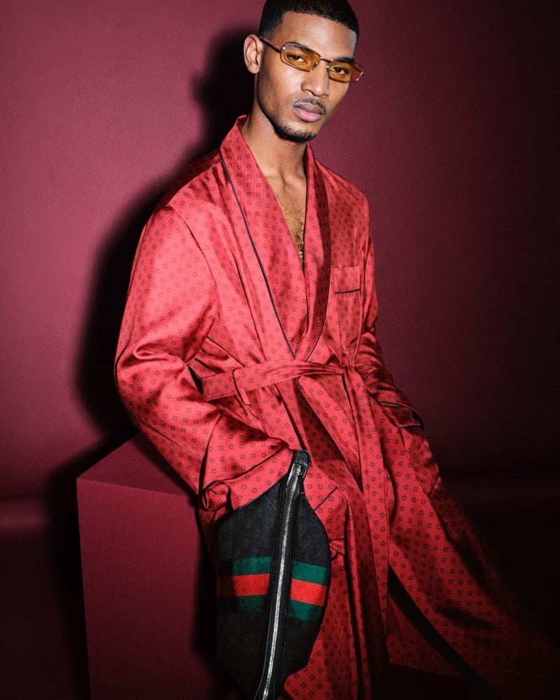 Reece Nelson seated on a burgundy plinth in a red silk dressing robe printed with a small dark diamond motif, belted at the waist, an oversized black GG canvas pouch with the Web stripe and chunky zipper resting across his lap, amber-tinted rectangular sunglasses and a short beard