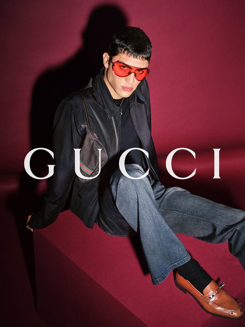 Roman Stubrin reclining on a deep burgundy plinth in a black leather zip jacket, gray flared trousers, and tan leather Horsebit loafers, a small GG-monogram canvas bag with Web stripe behind the shoulder, oval red-tinted sunglasses against a burgundy backdrop printed with the white Gucci logo