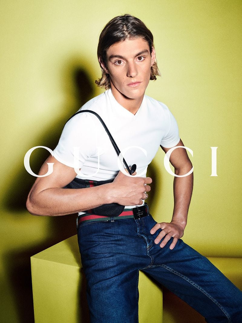 Vinicius Vieira leaning back on a yellow plinth in a fitted white short-sleeve V-neck tee and dark wash jeans cinched with the red-and-green Web stripe belt, a small GG-monogram canvas shoulder bag held against the torso against a chartreuse backdrop printed with the white Gucci logo