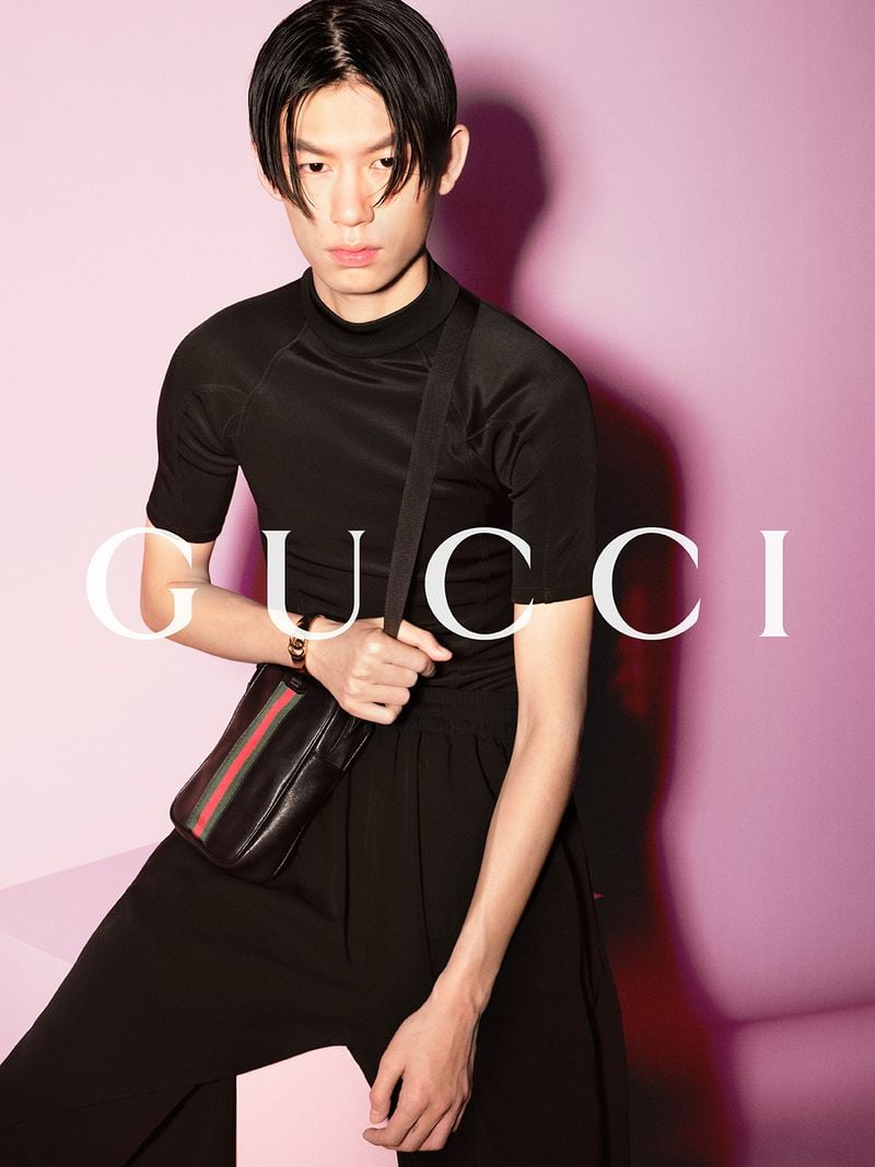 Yiming Ren against a soft pink backdrop in a fitted black short-sleeve mock-neck top and fluid black wide-leg trousers, a compact black leather Web-stripe shoulder bag pulled across the body, dark hair sweeping over the forehead