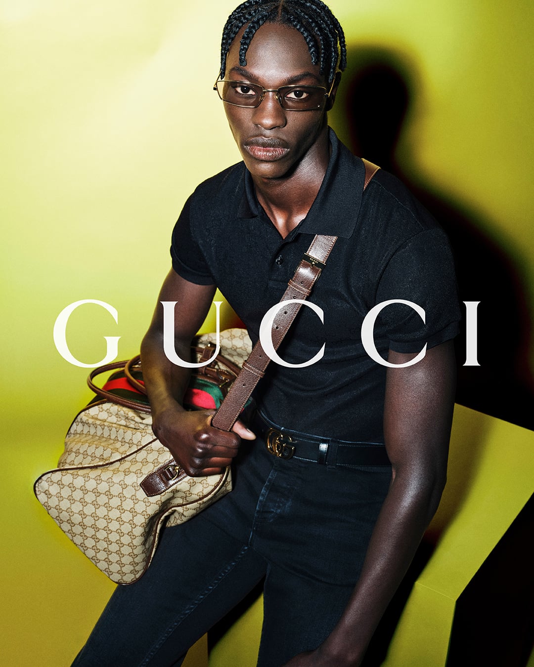 Close portrait of Abdou Thior against a chartreuse backdrop printed with the white Gucci logo, wearing a black short-sleeve polo and the black leather GG-buckle belt, an oversized GG-monogram canvas weekender with the red-and-green Web stripe held at his side, slim rimless sunglasses
